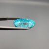 Close-up photograph of a 14.32-carat natural greenish-blue spiderweb Persian turquoise oval cabochon held in stainless steel tweezers against a neutral grey background, featuring intricate matrix patches and metallic pyrite inclusions.