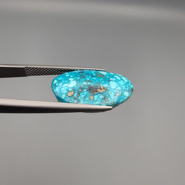 Close-up photograph of a 14.32-carat natural greenish-blue spiderweb Persian turquoise oval cabochon held in stainless steel tweezers against a neutral grey background, featuring intricate matrix patches and metallic pyrite inclusions.