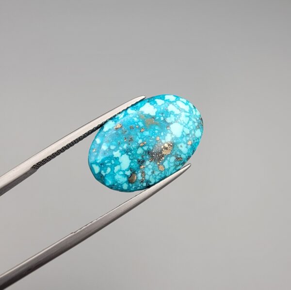 Close-up photograph of a 14.32-carat natural greenish-blue spiderweb Persian turquoise oval cabochon held in stainless steel tweezers against a neutral grey background, featuring intricate matrix patches and metallic pyrite inclusions.