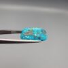 Close-up photograph of a 14.32-carat natural greenish-blue spiderweb Persian turquoise oval cabochon held in stainless steel tweezers against a neutral grey background, featuring intricate matrix patches and metallic pyrite inclusions.