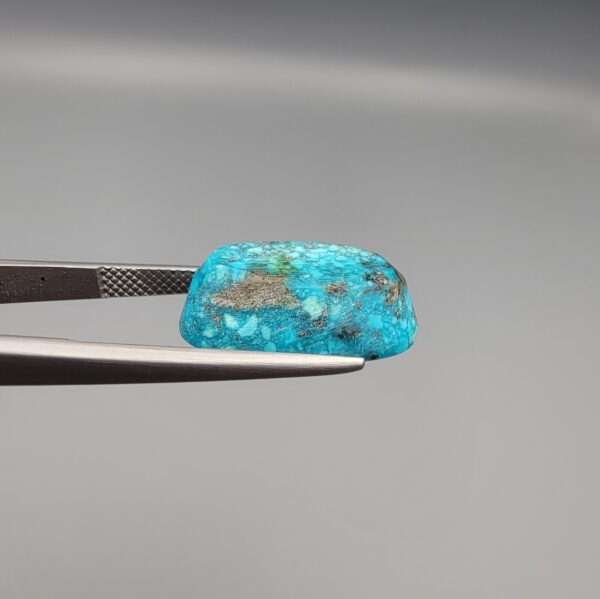 Close-up photograph of a 14.32-carat natural greenish-blue spiderweb Persian turquoise oval cabochon held in stainless steel tweezers against a neutral grey background, featuring intricate matrix patches and metallic pyrite inclusions.