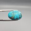Close-up photograph of a 14.32-carat natural greenish-blue spiderweb Persian turquoise oval cabochon held in stainless steel tweezers against a neutral grey background, featuring intricate matrix patches and metallic pyrite inclusions.