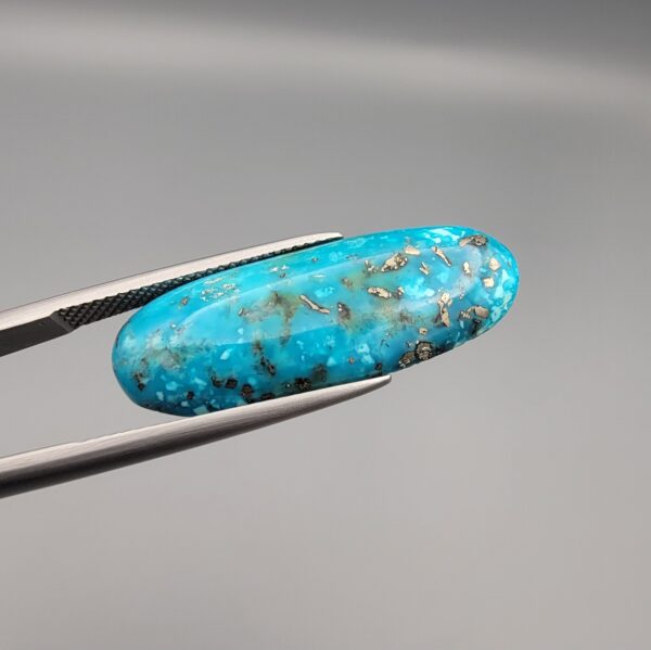 Close-up photograph of a 16.64-carat natural bright blue Persian turquoise elongated oval cabochon held in stainless steel tweezers against a neutral grey background, showcasing vibrant blue saturation with scattered metallic pyrite flecks on the face and a dense pyrite-rich reverse.