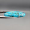 Close-up photograph of a 16.64-carat natural bright blue Persian turquoise elongated oval cabochon held in stainless steel tweezers against a neutral grey background, showcasing vibrant blue saturation with scattered metallic pyrite flecks on the face and a dense pyrite-rich reverse.