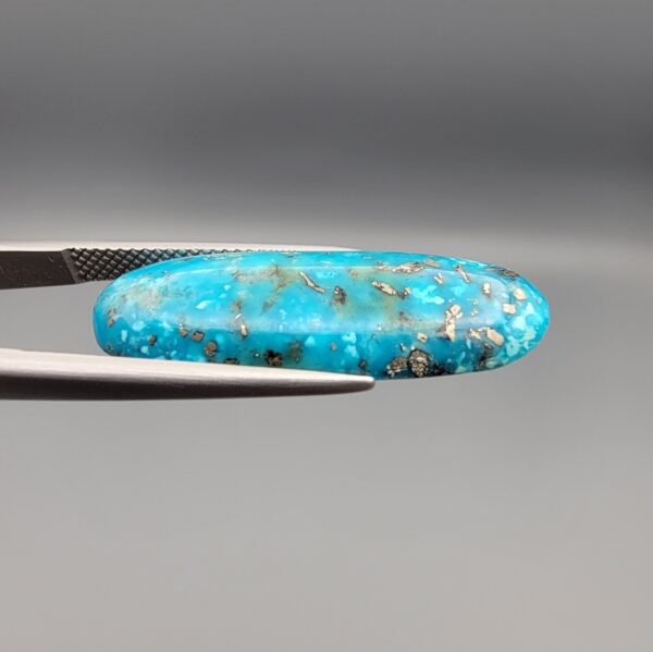 Close-up photograph of a 16.64-carat natural bright blue Persian turquoise elongated oval cabochon held in stainless steel tweezers against a neutral grey background, showcasing vibrant blue saturation with scattered metallic pyrite flecks on the face and a dense pyrite-rich reverse.