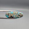 Close-up photograph of a 16.64-carat natural bright blue Persian turquoise elongated oval cabochon held in stainless steel tweezers against a neutral grey background, showcasing vibrant blue saturation with scattered metallic pyrite flecks on the face and a dense pyrite-rich reverse.
