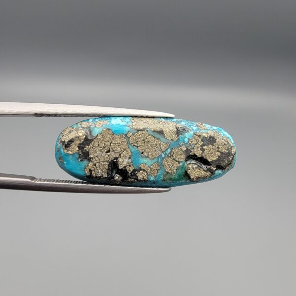 Close-up photograph of a 16.64-carat natural bright blue Persian turquoise elongated oval cabochon held in stainless steel tweezers against a neutral grey background, showcasing vibrant blue saturation with scattered metallic pyrite flecks on the face and a dense pyrite-rich reverse.