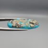 Close-up photograph of a 16.64-carat natural bright blue Persian turquoise elongated oval cabochon held in stainless steel tweezers against a neutral grey background, showcasing vibrant blue saturation with scattered metallic pyrite flecks on the face and a dense pyrite-rich reverse.