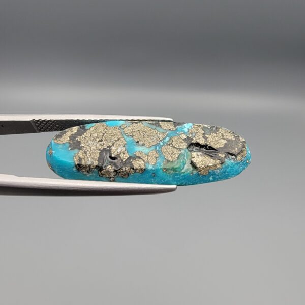Close-up photograph of a 16.64-carat natural bright blue Persian turquoise elongated oval cabochon held in stainless steel tweezers against a neutral grey background, showcasing vibrant blue saturation with scattered metallic pyrite flecks on the face and a dense pyrite-rich reverse.