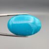 Close-up photograph of a 39.68-carat natural domed bright blue Sleeping Beauty turquoise (Razaki Feroza) oval cabochon held in tweezers against a neutral background, showcasing a speckled reverse side with visible texture and a light-colored host matrix inclusion.