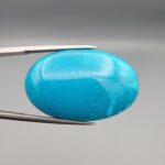 Close-up photograph of a 39.68-carat natural domed bright blue Sleeping Beauty turquoise (Razaki Feroza) oval cabochon held in tweezers against a neutral background, showcasing a speckled reverse side with visible texture and a light-colored host matrix inclusion.