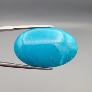 Close-up photograph of a 39.68-carat natural domed bright blue Sleeping Beauty turquoise (Razaki Feroza) oval cabochon held in tweezers against a neutral background, showcasing a speckled reverse side with visible texture and a light-colored host matrix inclusion.