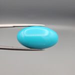Two-part macro photograph of an 18.01-carat natural bright blue Sleeping Beauty turquoise (Razaki Feroza) oval cabochon held in tweezers, showcasing a vibrant, uniform polished front face and a reverse side with a delicate branching white host matrix pattern.