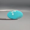 Two-part macro photograph of an 18.01-carat natural bright blue Sleeping Beauty turquoise (Razaki Feroza) oval cabochon held in tweezers, showcasing a vibrant, uniform polished front face and a reverse side with a delicate branching white host matrix pattern.
