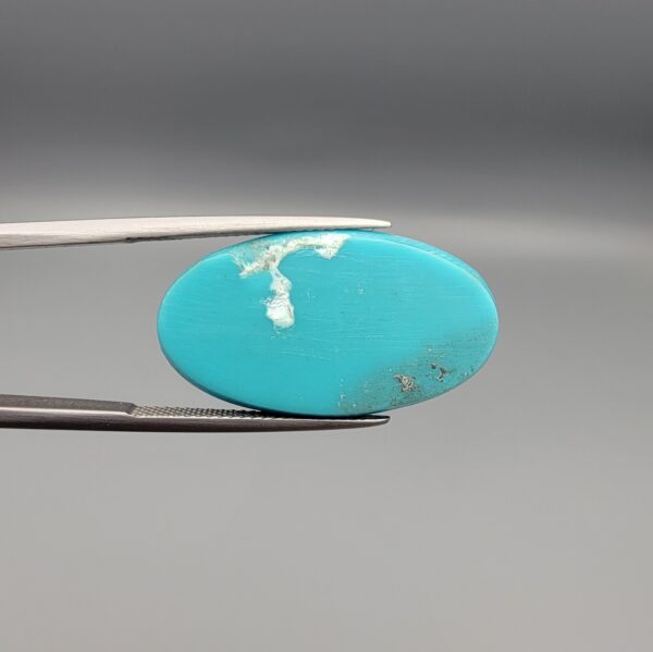 Two-part macro photograph of an 18.01-carat natural bright blue Sleeping Beauty turquoise (Razaki Feroza) oval cabochon held in tweezers, showcasing a vibrant, uniform polished front face and a reverse side with a delicate branching white host matrix pattern.