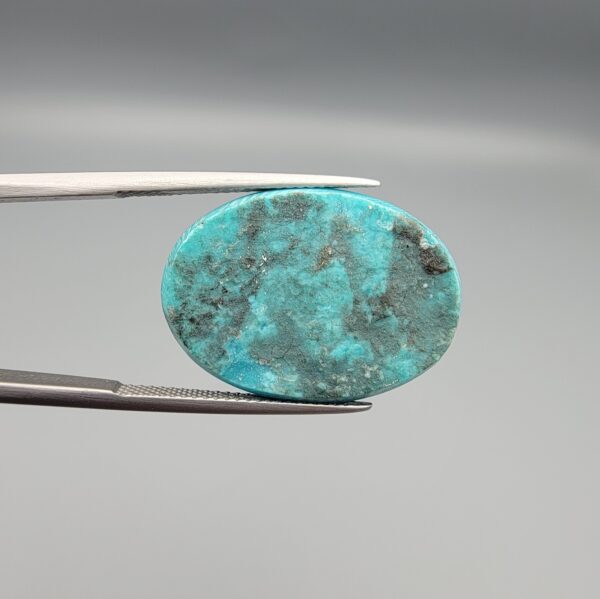 Multi-angle macro photographs of a 20.57-carat natural vibrant sky-blue Sleeping Beauty turquoise (Razaki Feroza) oval cabochon held in tweezers, showcasing a brilliantly polished front face and a reverse surface densely patterned with dark, mottled matrix inclusions.