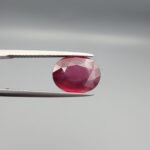 Macro photograph of a 4.27-carat natural oval-cut Rhodolite garnet from Madagascar held securely in metal tweezers, displaying deep raspberry-red coloration and precise facet symmetry.
