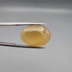 Multi-angle macro photographs of a 16.65-carat natural yellow chalcedony agate (Zard Aqeeq) oval cabochon from Iran held in metal tweezers, highlighting a smooth domed profile, uniform honey-yellow translucency, and a high-gloss reflective surface.