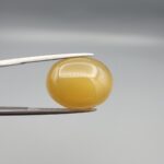 Multi-angle macro photographs of a 15.98-carat natural warm honey yellow chalcedony agate (Shehedi Zard Aqeeq) oval cabochon from Iran held in metal tweezers, showcasing a smooth domed profile, rich honey-colored translucency, and a mirror-like finish.