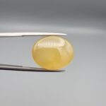 Multi-angle macro photographs of a 19.69-carat natural yellow chalcedony agate (Zard Aqeeq) oval cabochon from Iran held in metal tweezers, showcasing a smooth, domed surface, uniform honey-yellow translucency, and a high-gloss finish.