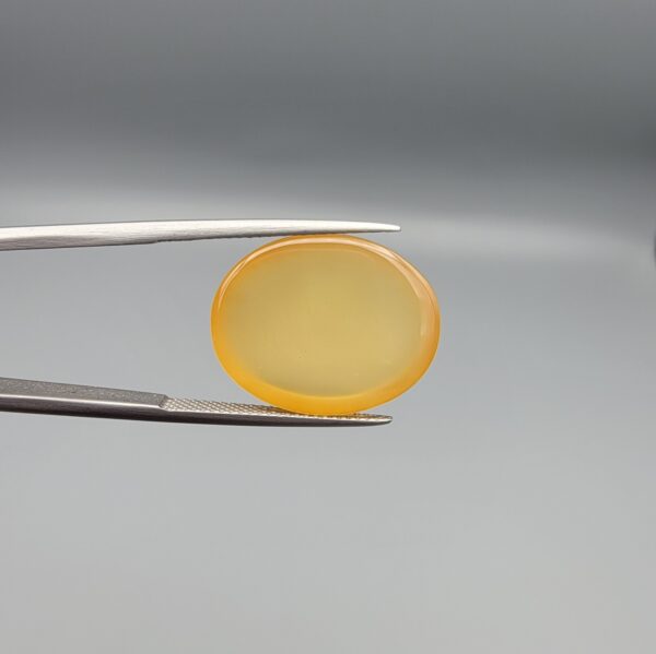 Multi-angle macro photographs of a 19.69-carat natural yellow chalcedony agate (Zard Aqeeq) oval cabochon from Iran held in metal tweezers, showcasing a smooth, domed surface, uniform honey-yellow translucency, and a high-gloss finish.