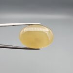 Multi-angle macro photographs of a 27.49-carat natural yellow chalcedony agate (Zard Aqeeq) oval cabochon from Iran held in metal tweezers, showcasing a smooth, domed profile, uniform honey-yellow translucency, and a high-gloss reflective surface.