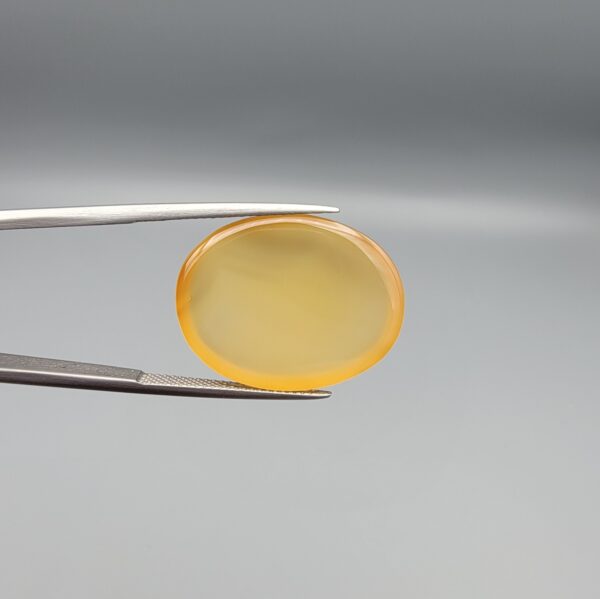 Multi-angle macro photographs of a 26.29-carat natural yellow chalcedony agate (Zard Aqeeq) oval cabochon from Iran held in metal tweezers, showcasing a smooth, domed profile, uniform honey-yellow translucency, and a high-gloss reflective surface.