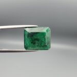 Macro photograph of a 6.46-carat natural emerald (Panna / Zamrud) from the Swat Valley in Pakistan held in metal tweezers, showcasing an emerald cut, a rich green body, and distinctive natural dark mineral dispersions across the stone.