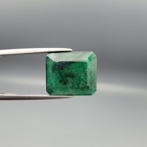 Macro photograph of a 6.46-carat natural emerald (Panna / Zamrud) from the Swat Valley in Pakistan held in metal tweezers, showcasing an emerald cut, a rich green body, and distinctive natural dark mineral dispersions across the stone.