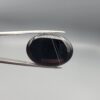 Multi-angle macro photographs of a 27.12-carat natural black banded agate (Sulemani Aqeeq) oval cabochon held in metal tweezers and fingertips, showcasing an intense deep black mirror finish crossed by a single fine, elegant curving white stripe across the face.