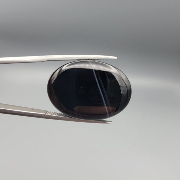 Multi-angle macro photographs of a 27.12-carat natural black banded agate (Sulemani Aqeeq) oval cabochon held in metal tweezers and fingertips, showcasing an intense deep black mirror finish crossed by a single fine, elegant curving white stripe across the face.