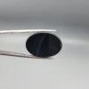 Multi-angle macro photographs of a 27.12-carat natural black banded agate (Sulemani Aqeeq) oval cabochon held in metal tweezers and fingertips, showcasing an intense deep black mirror finish crossed by a single fine, elegant curving white stripe across the face.