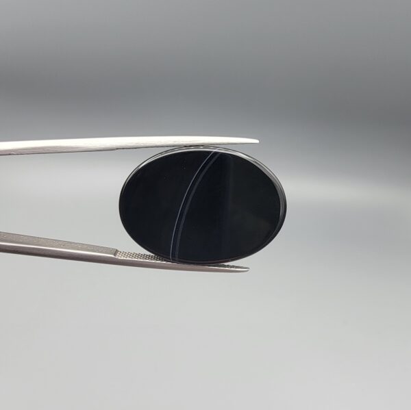 Multi-angle macro photographs of a 27.12-carat natural black banded agate (Sulemani Aqeeq) oval cabochon held in metal tweezers and fingertips, showcasing an intense deep black mirror finish crossed by a single fine, elegant curving white stripe across the face.