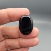 Multi-angle macro photographs of a 27.12-carat natural black banded agate (Sulemani Aqeeq) oval cabochon held in metal tweezers and fingertips, showcasing an intense deep black mirror finish crossed by a single fine, elegant curving white stripe across the face.