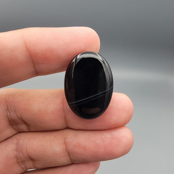 Multi-angle macro photographs of a 27.12-carat natural black banded agate (Sulemani Aqeeq) oval cabochon held in metal tweezers and fingertips, showcasing an intense deep black mirror finish crossed by a single fine, elegant curving white stripe across the face.