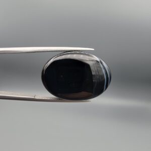 Multi-angle macro photographs of a 26.33-carat natural black banded agate (Sulemani Aqeeq) oval cabochon held in metal tweezers, showcasing a mirror-polished obsidian black surface accented with three distinct, parallel curving white stripes across its face.