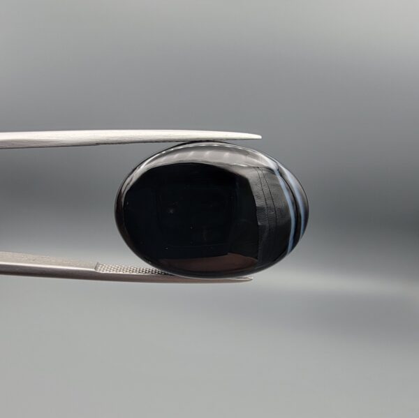 Multi-angle macro photographs of a 26.33-carat natural black banded agate (Sulemani Aqeeq) oval cabochon held in metal tweezers, showcasing a mirror-polished obsidian black surface accented with three distinct, parallel curving white stripes across its face.