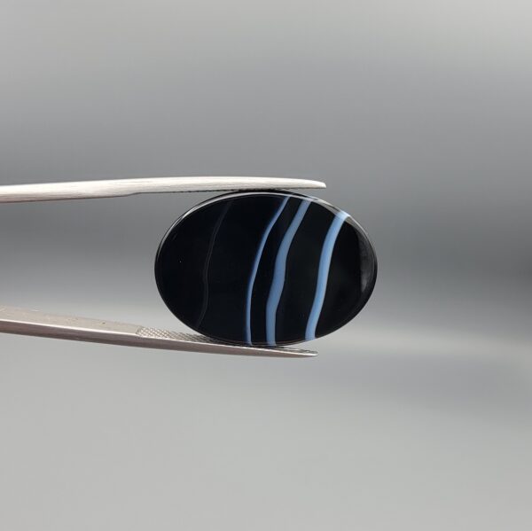 Multi-angle macro photographs of a 26.33-carat natural black banded agate (Sulemani Aqeeq) oval cabochon held in metal tweezers, showcasing a mirror-polished obsidian black surface accented with three distinct, parallel curving white stripes across its face.