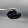Multi-angle macro photographs of a 26.88-carat natural black banded agate (Sulemani Aqeeq) oval cabochon held in metal tweezers, showcasing a high-gloss obsidian black dome with an intricate, wavy organic white veining pattern across the surface.