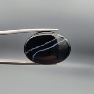 Multi-angle macro photographs of a 26.88-carat natural black banded agate (Sulemani Aqeeq) oval cabochon held in metal tweezers, showcasing a high-gloss obsidian black dome with an intricate, wavy organic white veining pattern across the surface.