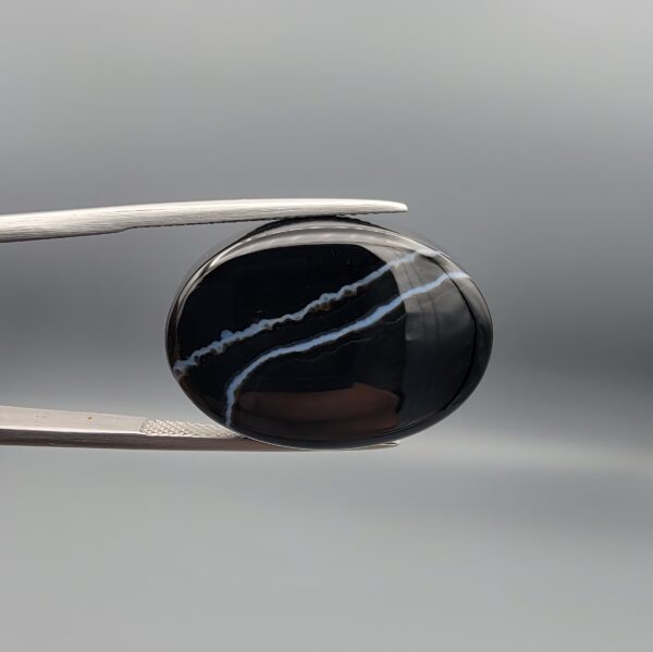 Multi-angle macro photographs of a 26.88-carat natural black banded agate (Sulemani Aqeeq) oval cabochon held in metal tweezers, showcasing a high-gloss obsidian black dome with an intricate, wavy organic white veining pattern across the surface.