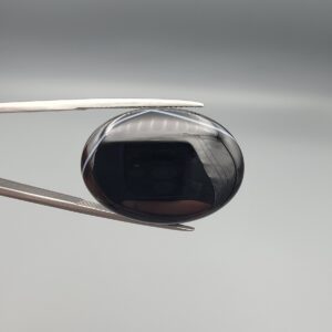 Multi-angle macro photographs of a 26.96-carat natural black banded agate (Sulemani Aqeeq) oval cabochon held in metal tweezers and fingertips, showcasing a mirror-polished obsidian black surface with sharp, angular chevron-style white banding lines across the face.