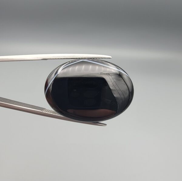 Multi-angle macro photographs of a 26.96-carat natural black banded agate (Sulemani Aqeeq) oval cabochon held in metal tweezers and fingertips, showcasing a mirror-polished obsidian black surface with sharp, angular chevron-style white banding lines across the face.
