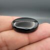 Multi-angle macro photographs of a 26.96-carat natural black banded agate (Sulemani Aqeeq) oval cabochon held in metal tweezers and fingertips, showcasing a mirror-polished obsidian black surface with sharp, angular chevron-style white banding lines across the face.