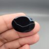 Multi-angle macro photographs of a 26.96-carat natural black banded agate (Sulemani Aqeeq) oval cabochon held in metal tweezers and fingertips, showcasing a mirror-polished obsidian black surface with sharp, angular chevron-style white banding lines across the face.