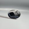 Multi-angle macro photographs of a 27.51-carat natural black banded agate (Sulemani Aqeeq) oval cabochon held in metal tweezers and fingertips, showcasing a mirror-polished deep obsidian black surface accented by fine, undulating white banding lines traversing across the polished surface.
