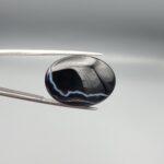 Multi-angle macro photographs of a 27.51-carat natural black banded agate (Sulemani Aqeeq) oval cabochon held in metal tweezers and fingertips, showcasing a mirror-polished deep obsidian black surface accented by fine, undulating white banding lines traversing across the polished surface.