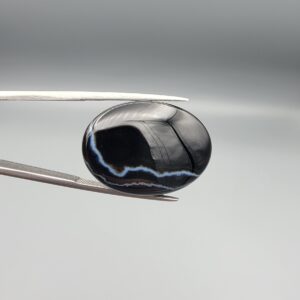 Multi-angle macro photographs of a 27.51-carat natural black banded agate (Sulemani Aqeeq) oval cabochon held in metal tweezers and fingertips, showcasing a mirror-polished deep obsidian black surface accented by fine, undulating white banding lines traversing across the polished surface.