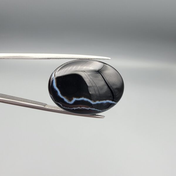 Multi-angle macro photographs of a 27.51-carat natural black banded agate (Sulemani Aqeeq) oval cabochon held in metal tweezers and fingertips, showcasing a mirror-polished deep obsidian black surface accented by fine, undulating white banding lines traversing across the polished surface.