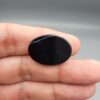 Multi-angle macro photographs of a 27.51-carat natural black banded agate (Sulemani Aqeeq) oval cabochon held in metal tweezers and fingertips, showcasing a mirror-polished deep obsidian black surface accented by fine, undulating white banding lines traversing across the polished surface.